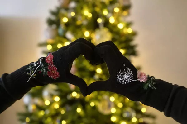 Behandschuhte Hände formen vor einem leuchtenden Weihnachtsbaum ein Herz.